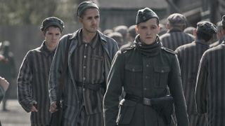 Jonah Hauer-King in a striped concentration camp uniform as Lali walks behind Jonas Nay in an SS uniform as Baretzki in The Tattooist of Auschwitz