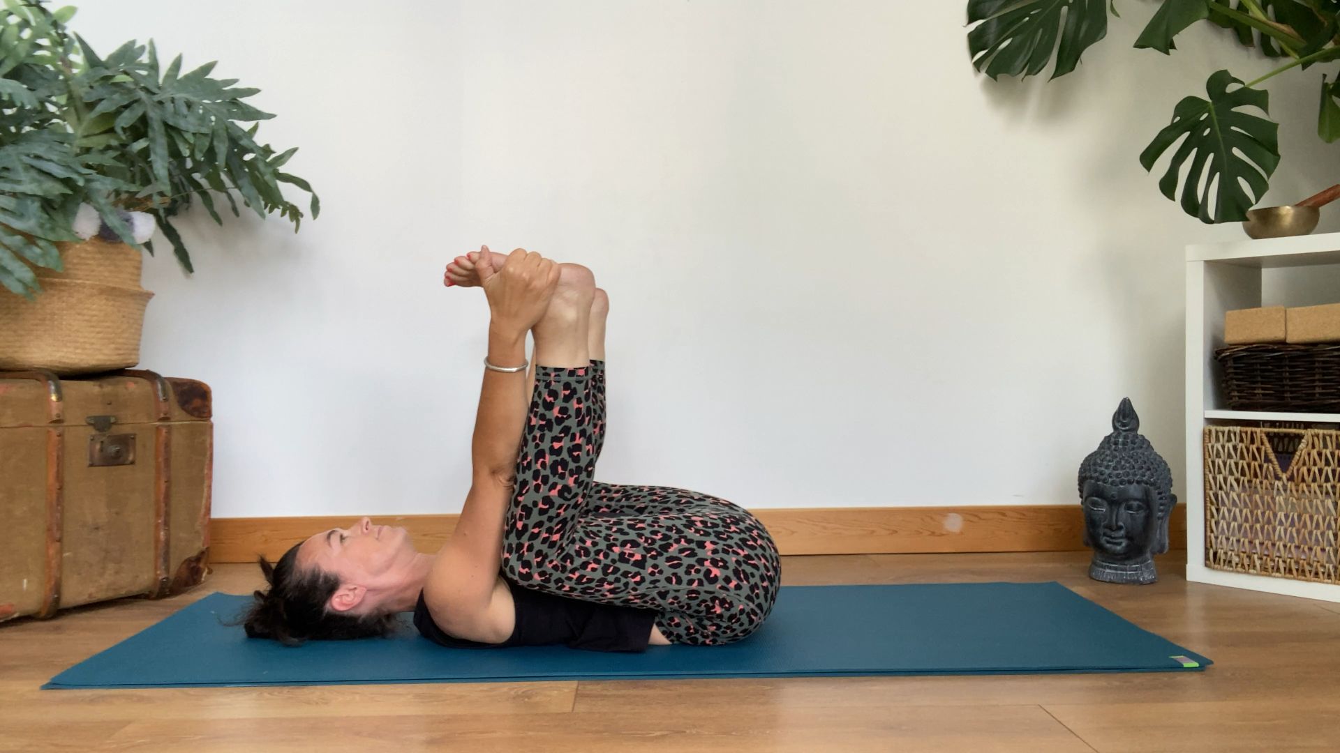 Louise Bartlett performs a happy baby pose at home on a yoga mat. She is lying on her back, with her knees bent and feet in the air, thighs close to her torso. She holds the soles of her feet in her hands.
