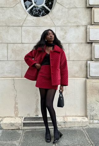 how to wear velvet is shown in a photo of a woman on the sidewalk wearing gold earrings, a black scarf, a quilted velvet jacket, a black v-neck sweater, a red quilted mini skirt, black tights, and black mary jane heels