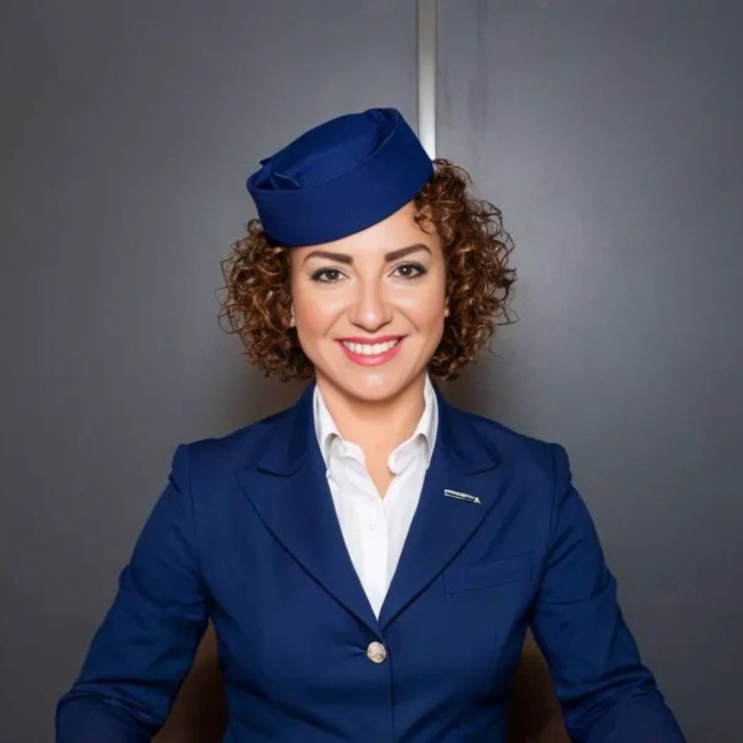 A cheerful headshot of Paula S Adams, a flight attendant with curly dark brown hair wearing a navy flight attendant uniform.