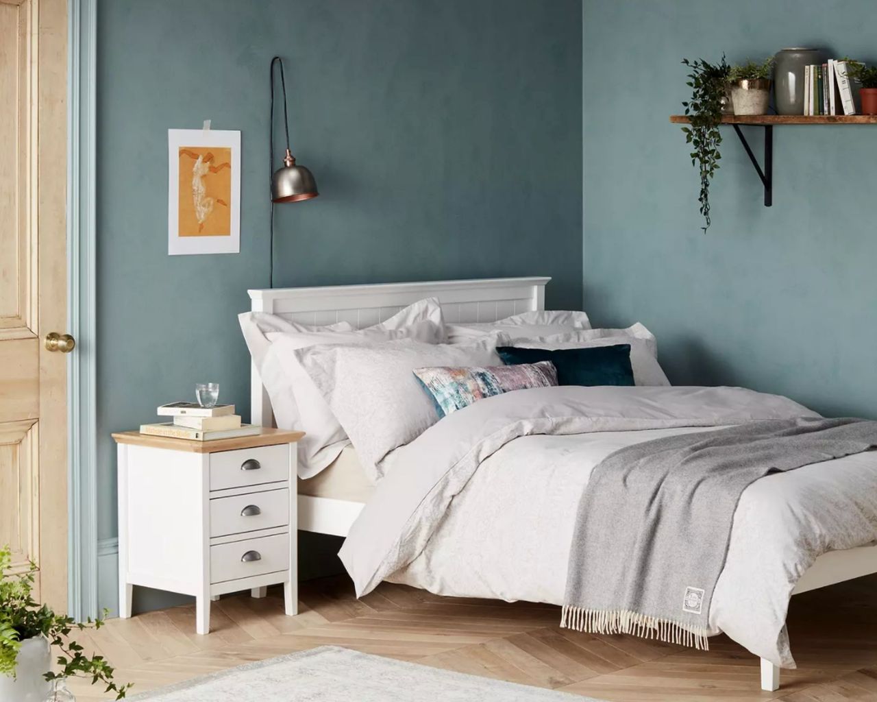 White wooden bed frame inside blue room pushed up against corner with wooden shelf, grey bedding and white bedside table