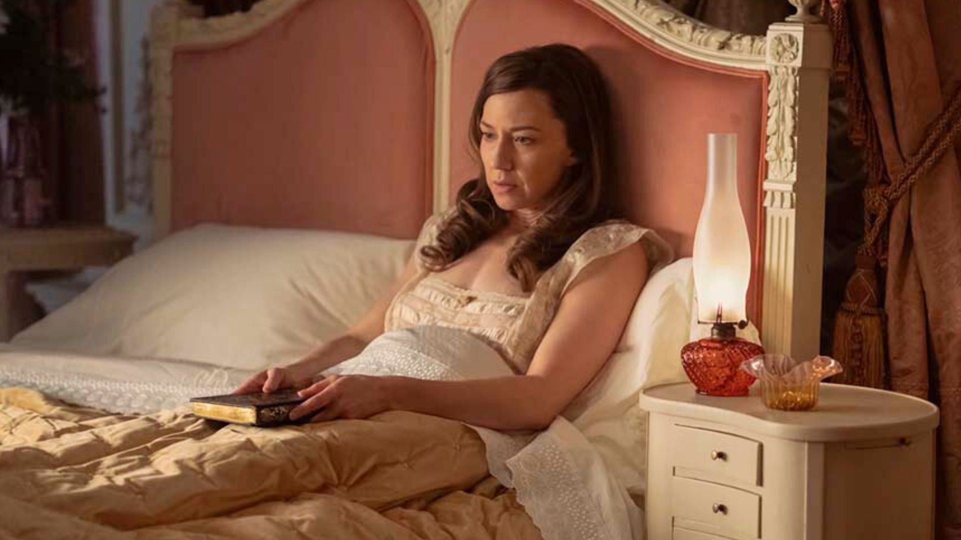 Bertha sits in bed looking distressed while holding a book