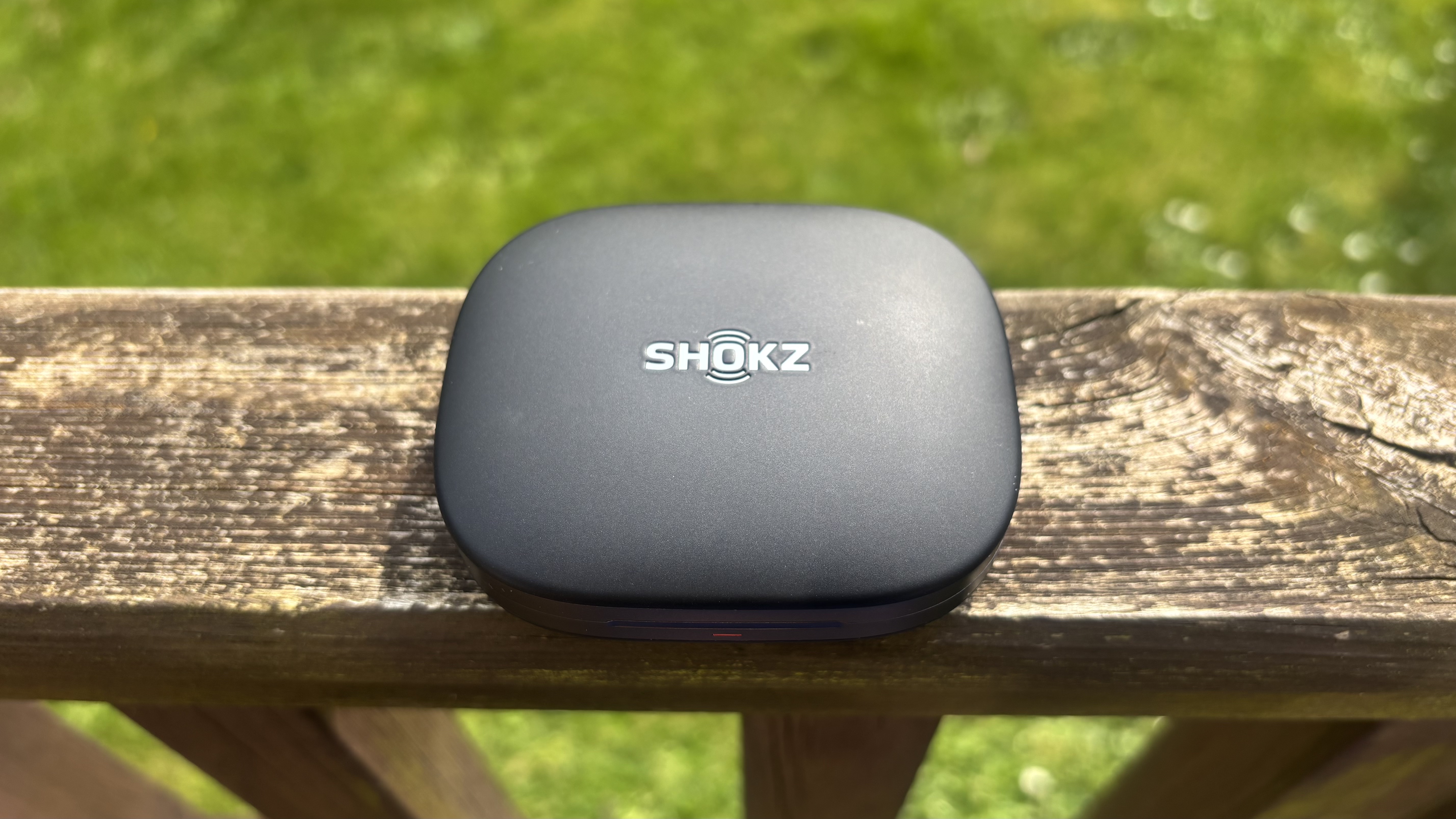 Shokz OpenFit Pro open earbuds charging case on a wooden bannister with grass in the background.