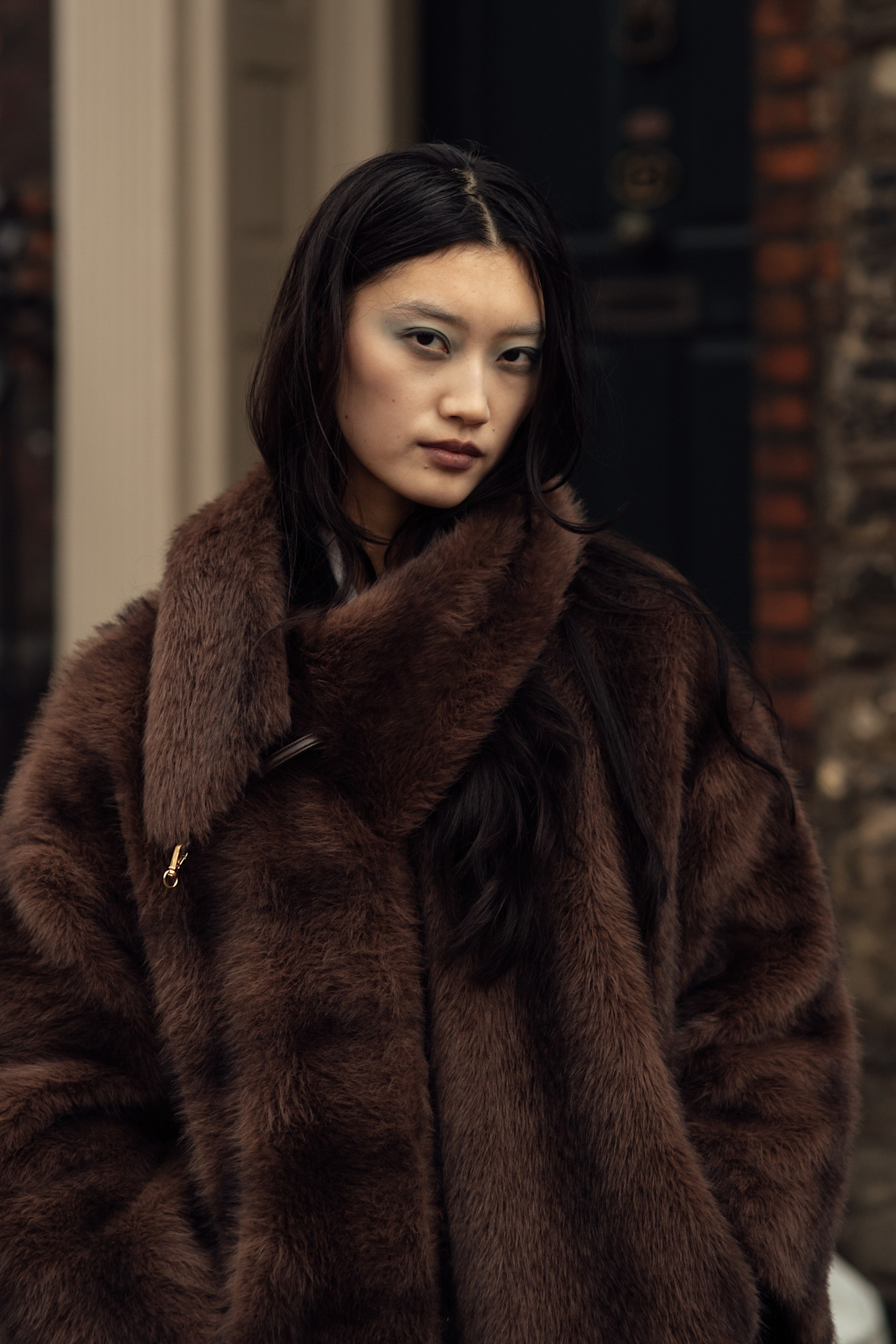 A woman wearing a faux fur brown coat, with hazy blue eyeshadow and brown lipstick