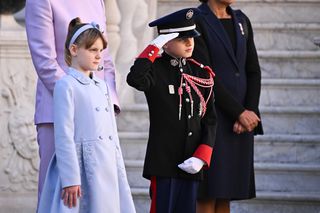 Prince Jacques of Monaco wearing a uniform and saluting while standing next to sister Princess Gabriella outside the palace