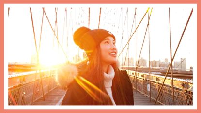 Woman in winter clothing smiling and looking up with sun in background on Brooklyn Bridge
