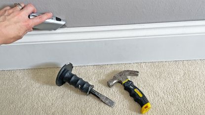 tools on carpeted floor in front of a white skirting board being removed by a hand holding a utility knife