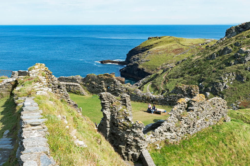 A series of fragmented stone ruins on green cliffsides overlooking a deep blue sea.