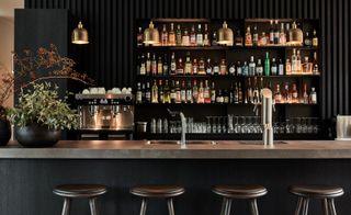 Interior view of the bar at Mauritzhof, Münster, Germany featuring a black and grey counter with a sink, bar stools, plants in curved pots, a coffee machine and a shelving unit on a black wall filled with glasses and bottles of drinks