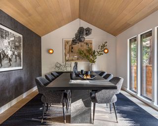 Dining room with vaulted ceiling, cladded in warming wood, large black marble dining table with upholstered dining chairs, modern hanging light fixture, blue rug on wooden floor, gray textured feature wall with artwork