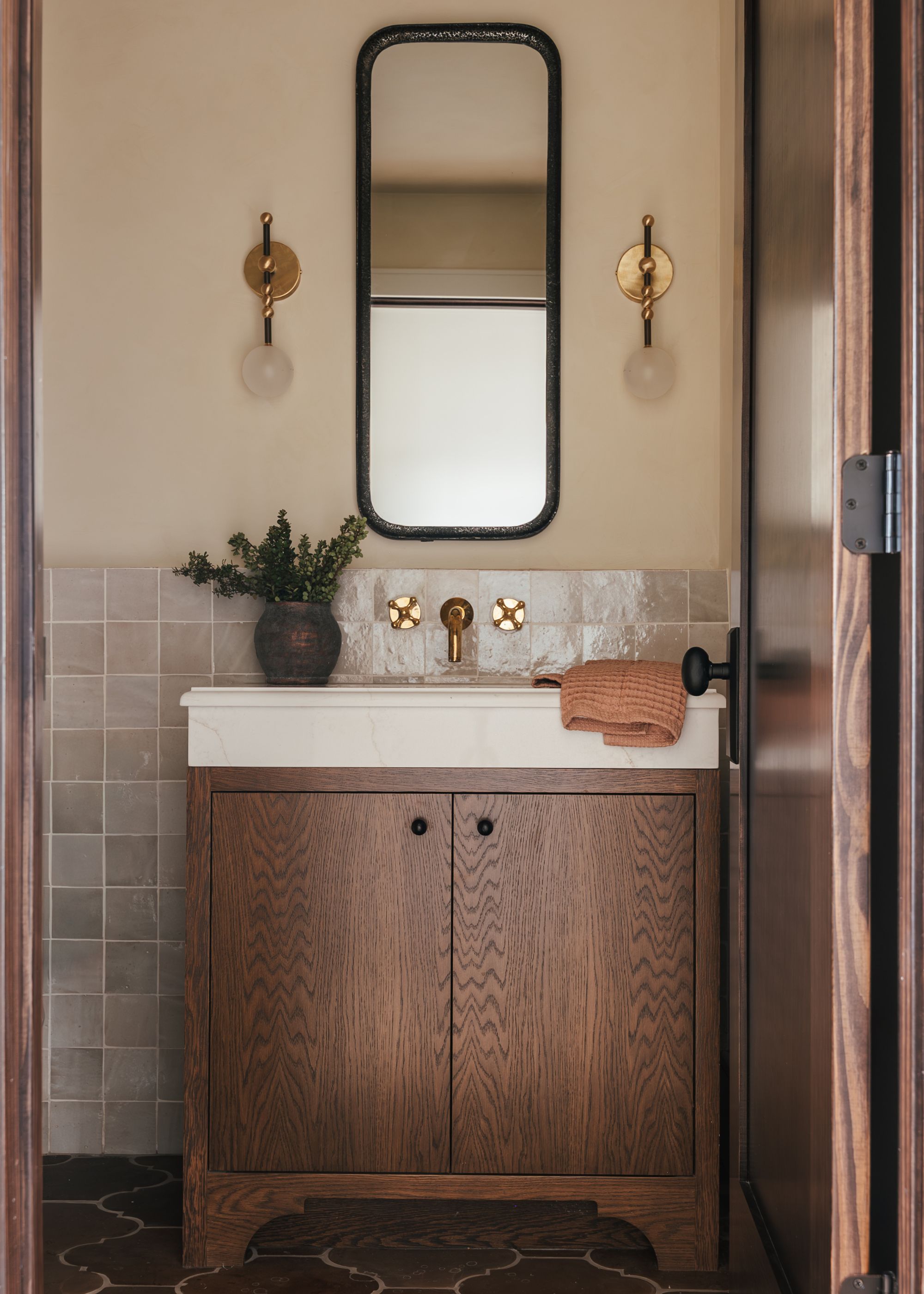 A small bathroom with wall sconces, a wall mirror, tiled flooring, and a vase of green stems on the sink