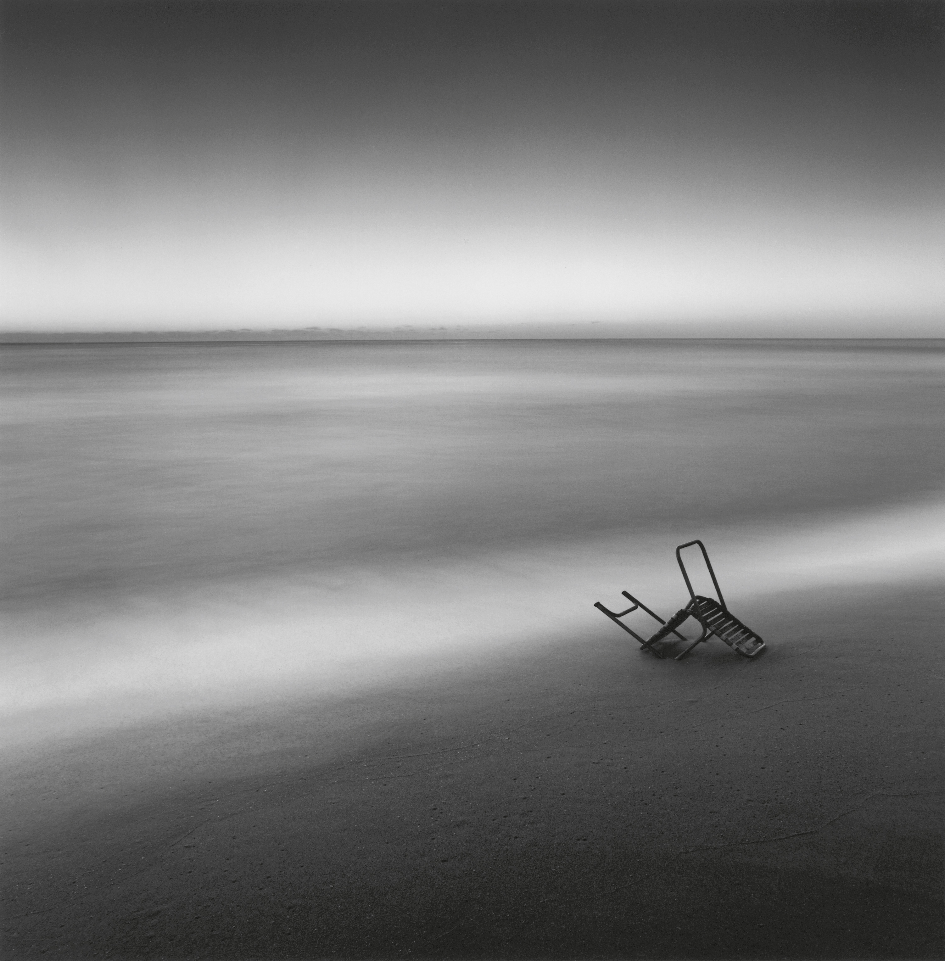 A lone, overturned metal chair rests on a smooth beach at the edge of a calm, blurred ocean under a gradient sky.