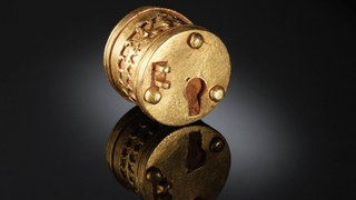 A small gold cylinder with rivets and a keyhole sits on a reflective black table