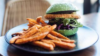 Crumbed tofu burger in a green bun with sweet potato fries sitting on plate on wooden table with wicker chair next to it