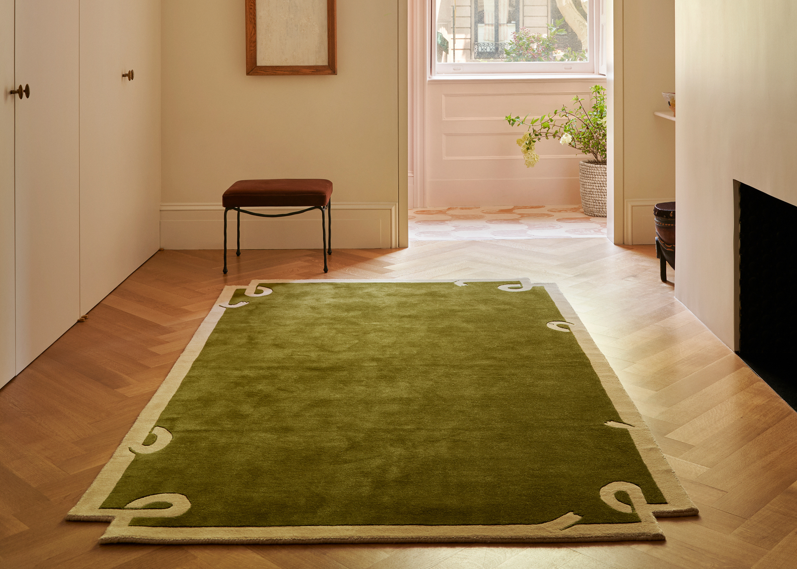 A room with wood flooring, a green rug with a cream frame, and a brown stool