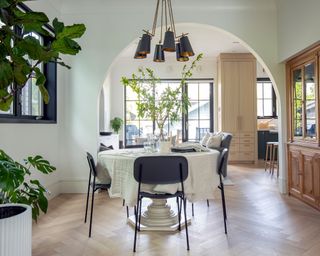 Modern dining room with round table, black chairs in center of room with wooden hutch visible to the right with indoor plants to the left
