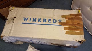 Damaged Winkbed Plus cardboard box, the sides are ripped up and 11 strips of brown tape are holding one side closed