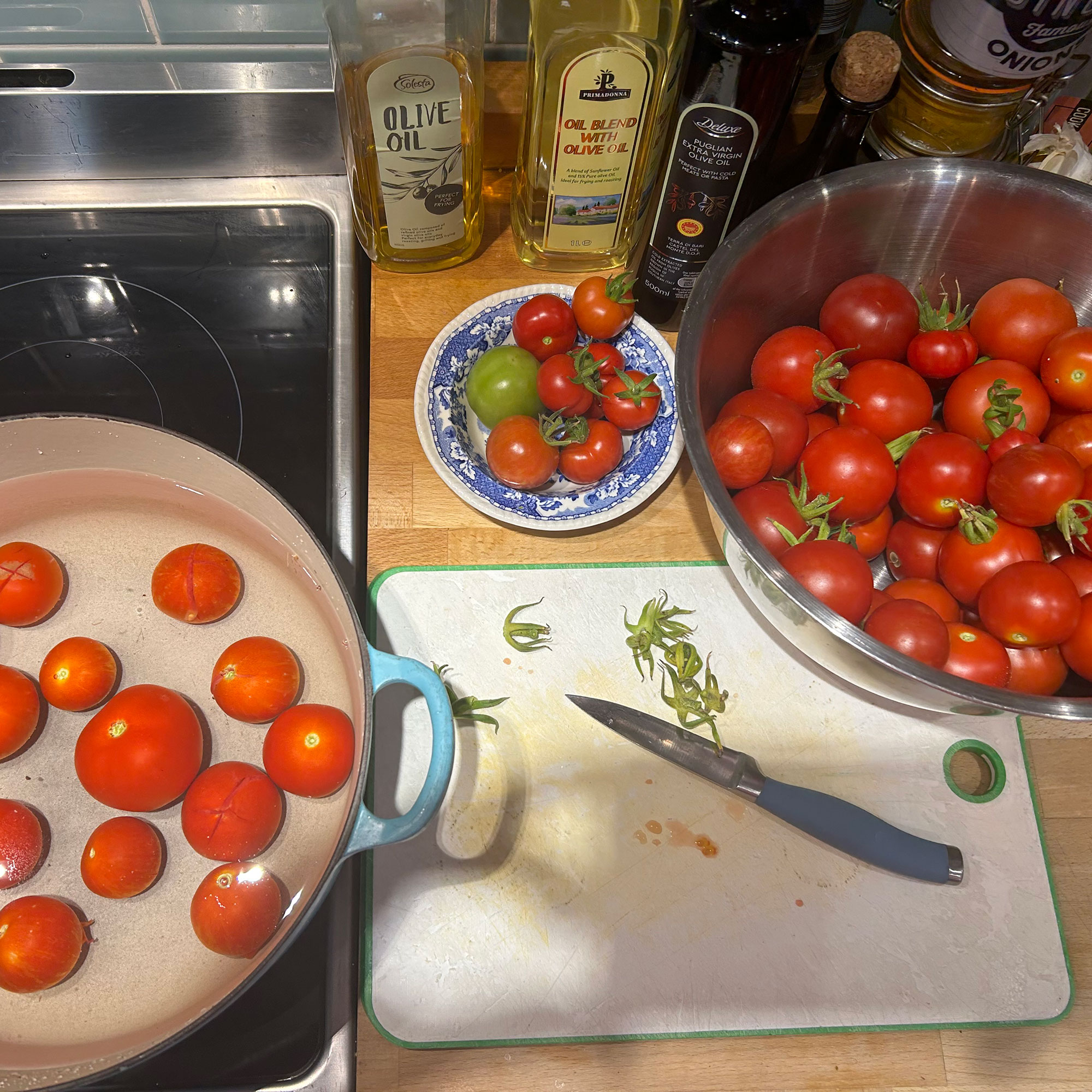 Kitchen worktop with chopping board viewed from above, with stainless steel bowl full of tomatoes on one side and casserole pit of tomatoes in water resting on hob on the other