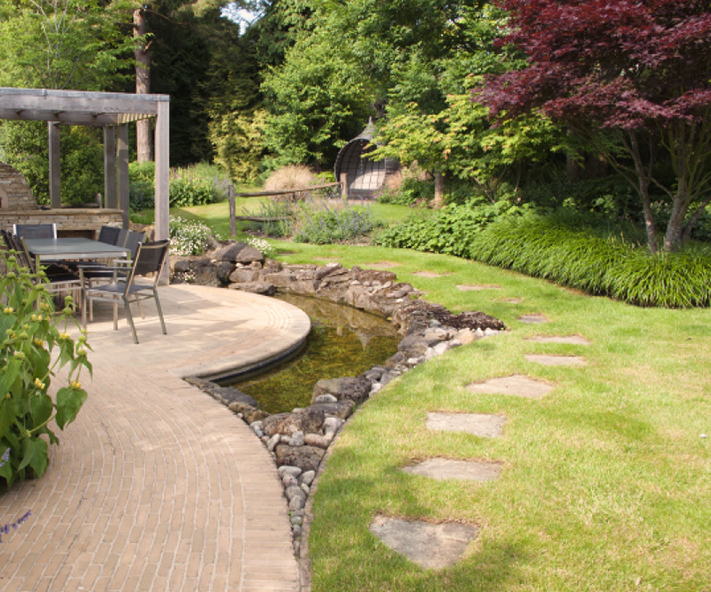 naturalistic water feature with stones, set alongside a patio area, with lawn on the other side and a stepping stone path, and a backdrop of trees