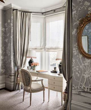 A white dressing table in a room with gray patterned wallpaper and traditional taupe drapes.
