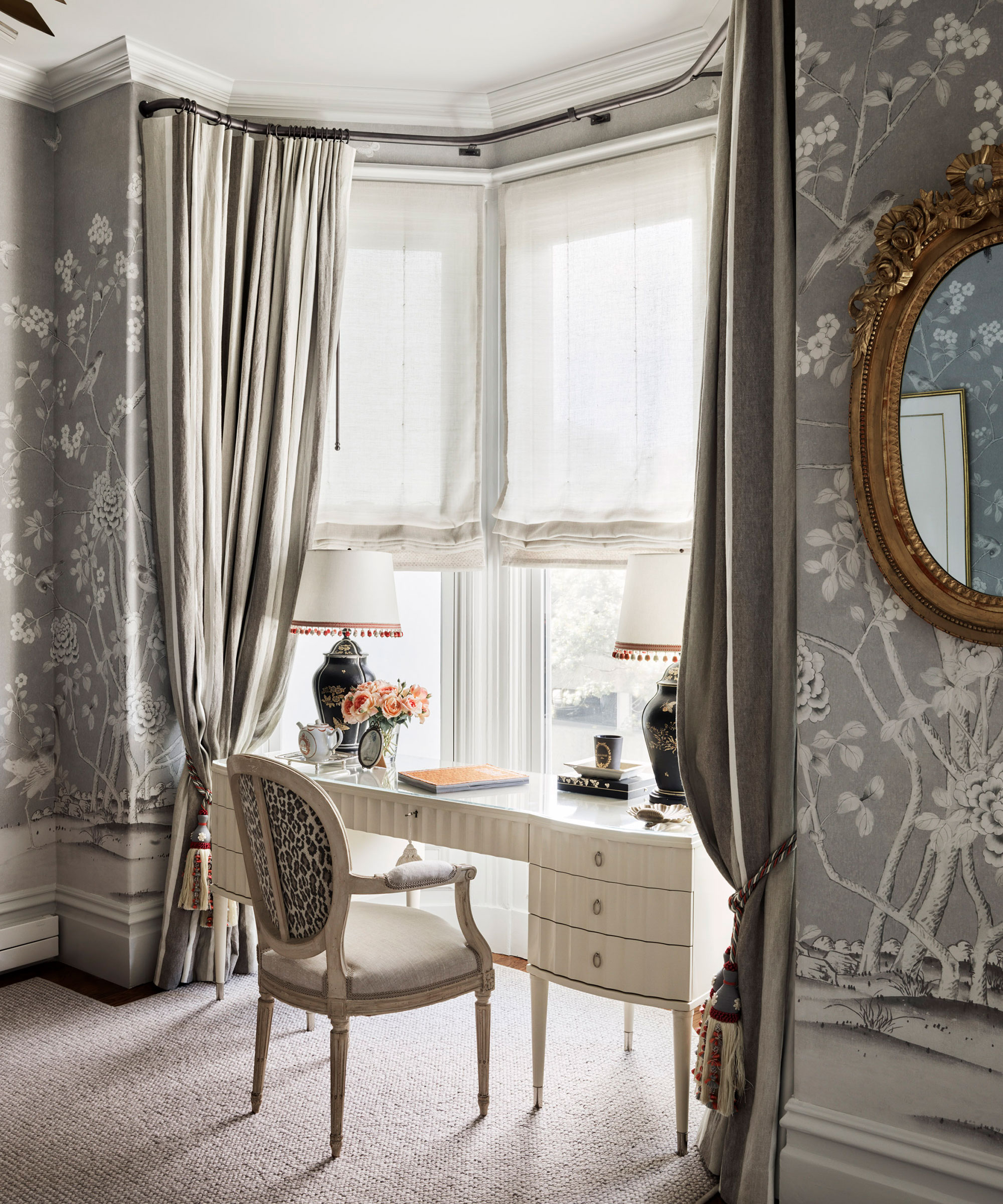A white dressing table in a room with gray patterned wallpaper and traditional taupe drapes.