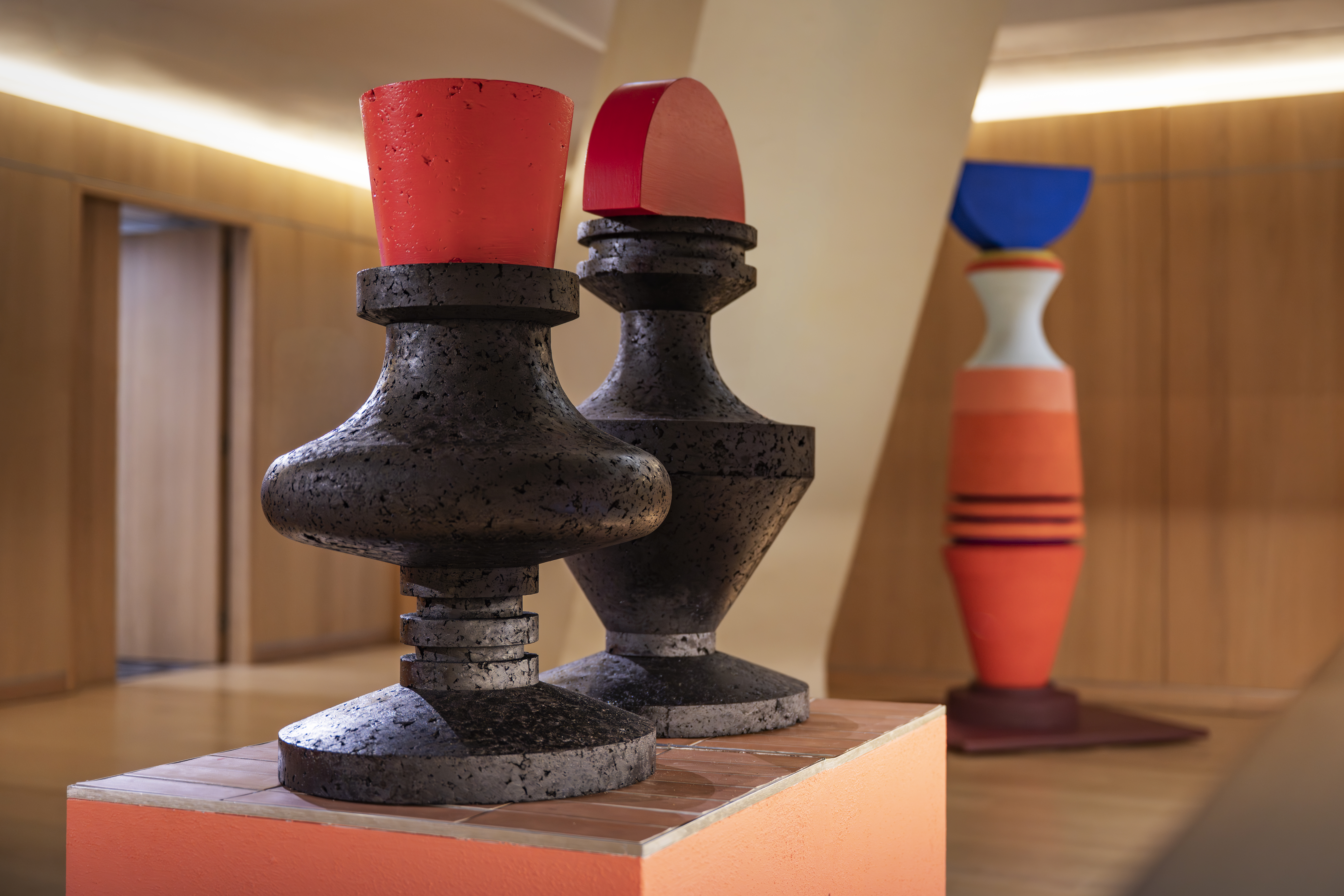 Three tall wooden sculptures with abstract, geometrical shapes, painted in vibrant hues of red, orange, and blue, sit in a wood-clad, brightly lit gallery.