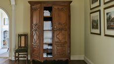 Dark wood linen closet in a neutral hallway with gallery wall, chair, and arched doorway