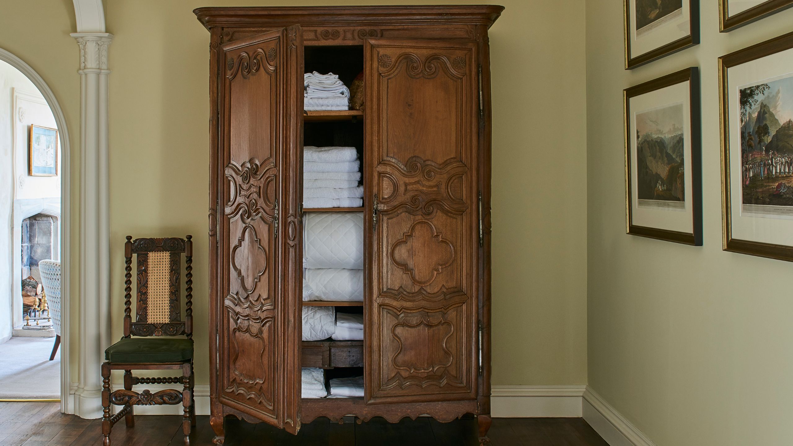 Dark wood linen closet in a neutral hallway with gallery wall, chair, and arched doorway