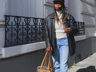 Fashion influencer @aude_julie on the street wearing a vintage-look, collared, black leather jacket with a turtleneck, blue faded jeans, burgundy heels, a Louis Vuitton handbag, and cap.