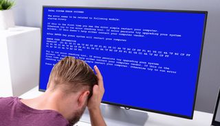 An old-fashioned Blue Screen of Death on a widescreen monitor while a user slams his palm onto his forehead.
