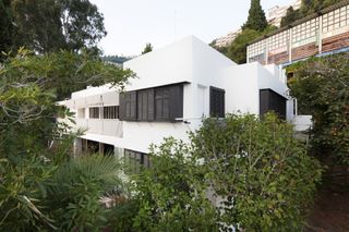 A double storey white villa surrounded by trees with houses on the hills behind it.