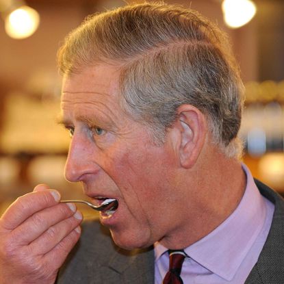 King Charles wears a gray suit and a lilac shirt and eats some food from a spoon