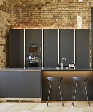 A kitchen with a black kitchen island with a silver faucet, two black bar stools, black cabinets with gold trims behind it, and exposed brown brick wall behind that