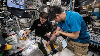 a Japanese man wearing a blue polo and khaki pants and an American woman wearing a black shirt and blue slacks work to draw and collect blood from themselves aboard a space station