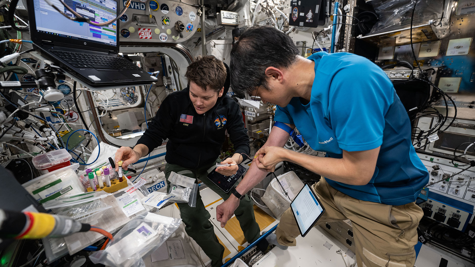 a Japanese man wearing a blue polo and khaki pants  and an American woman wearing a black shirt and blue slacks work to draw and collect blood from themselves aboard a space station