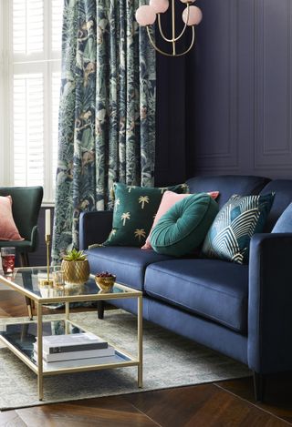 Luxe, dark living room with navy velvet couch, jungle inspired curtains, brass and glass coffee table, and teal and emerald scatter pillows.