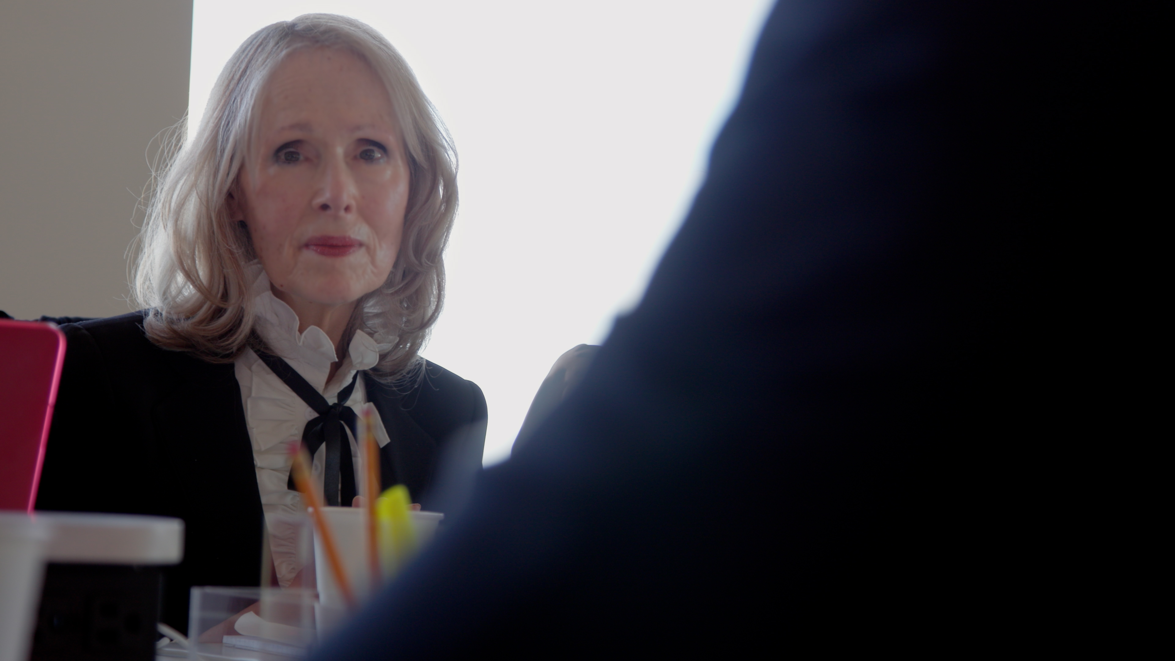 e. jean carroll wears a top with a bow around the neck and a blazer at a desk in a still from the documentary ask e jean