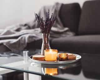 Glass coffee table with plate of candles and vase of purple flowers, with grey sofa in background