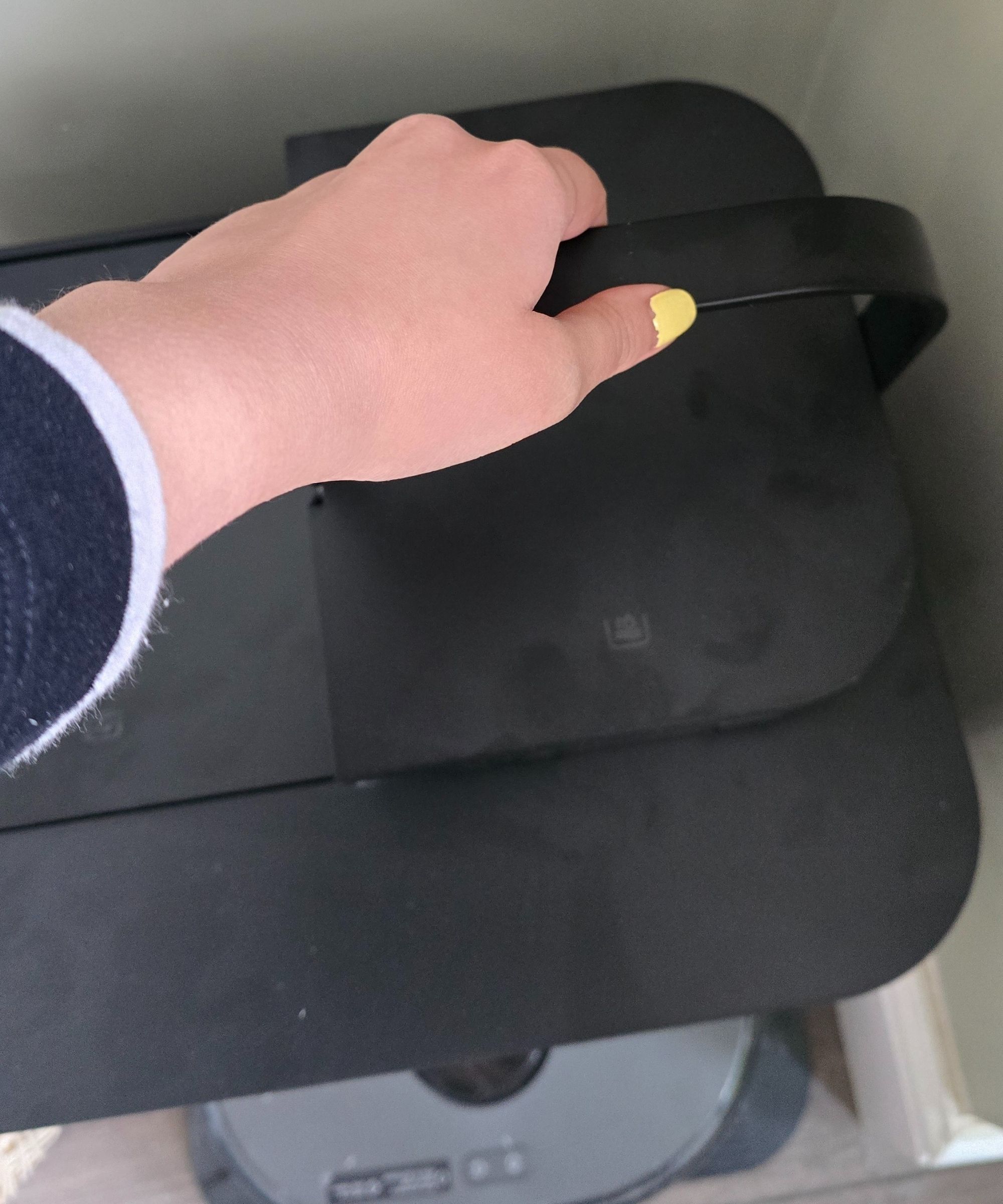 A hand with yellow nail varnish on the nails holding a black water tank, with a black robot vacuum and base underneath