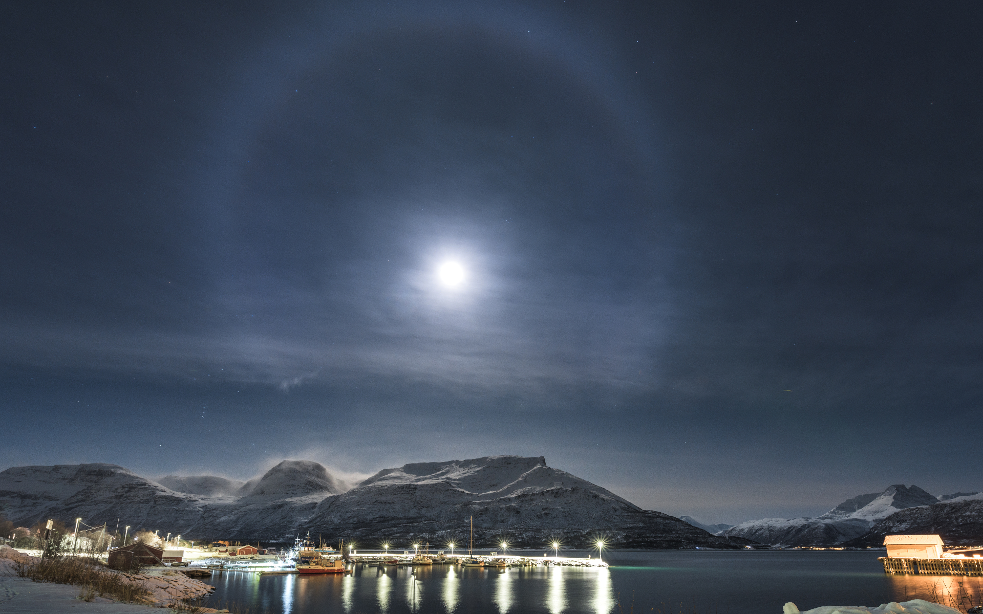 Halo around the moon which is located in the sky above snow-capped mountains and a large body of water below. Lights from a nearby town are reflected in the water.
