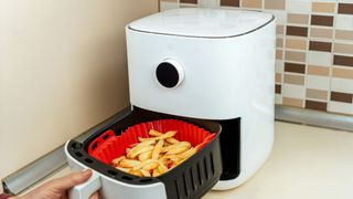 A white air fryer next to a wall and cupboard, with a slightly open basket