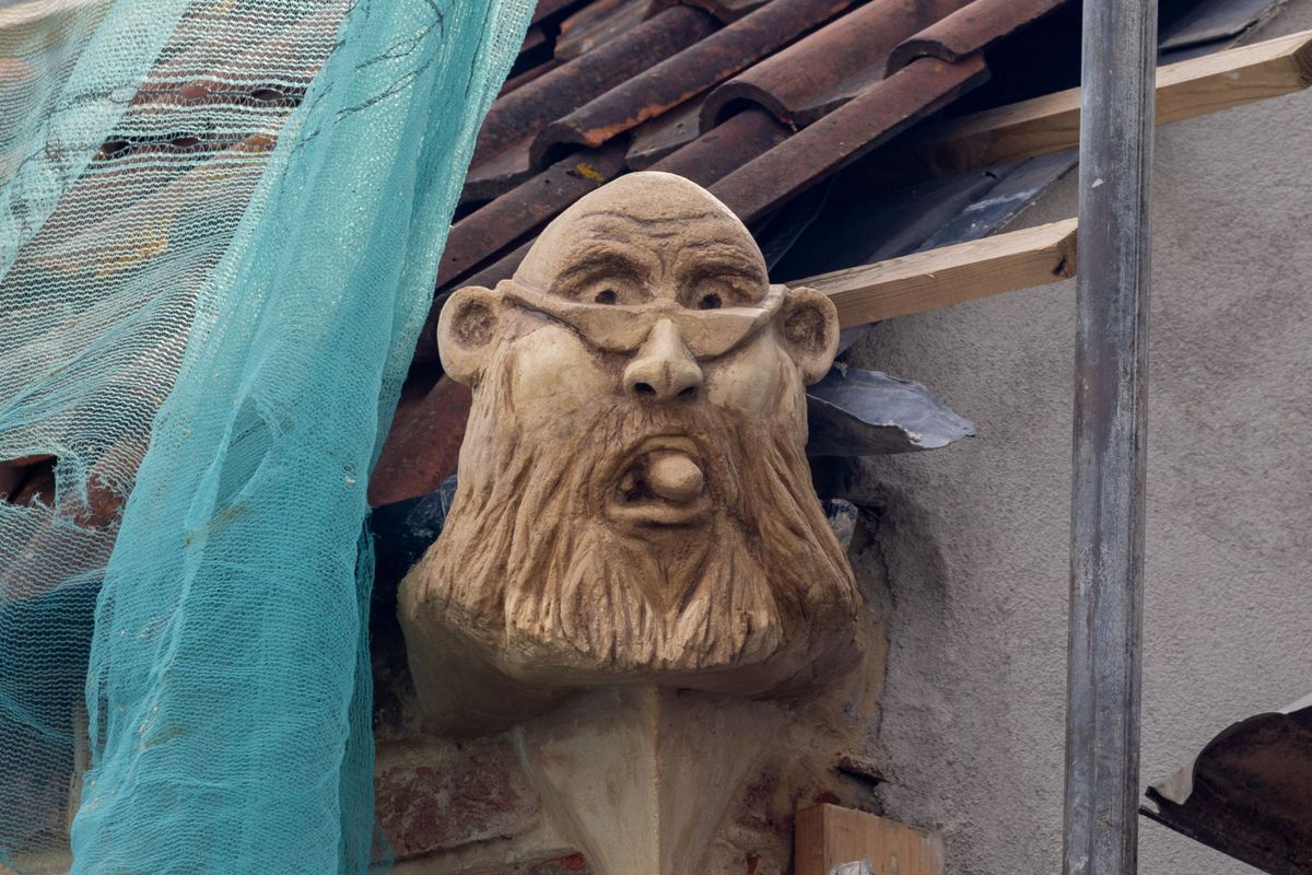 A stone grotesque on top of a building with platforming around it depicting a bald man with a beard and glasses