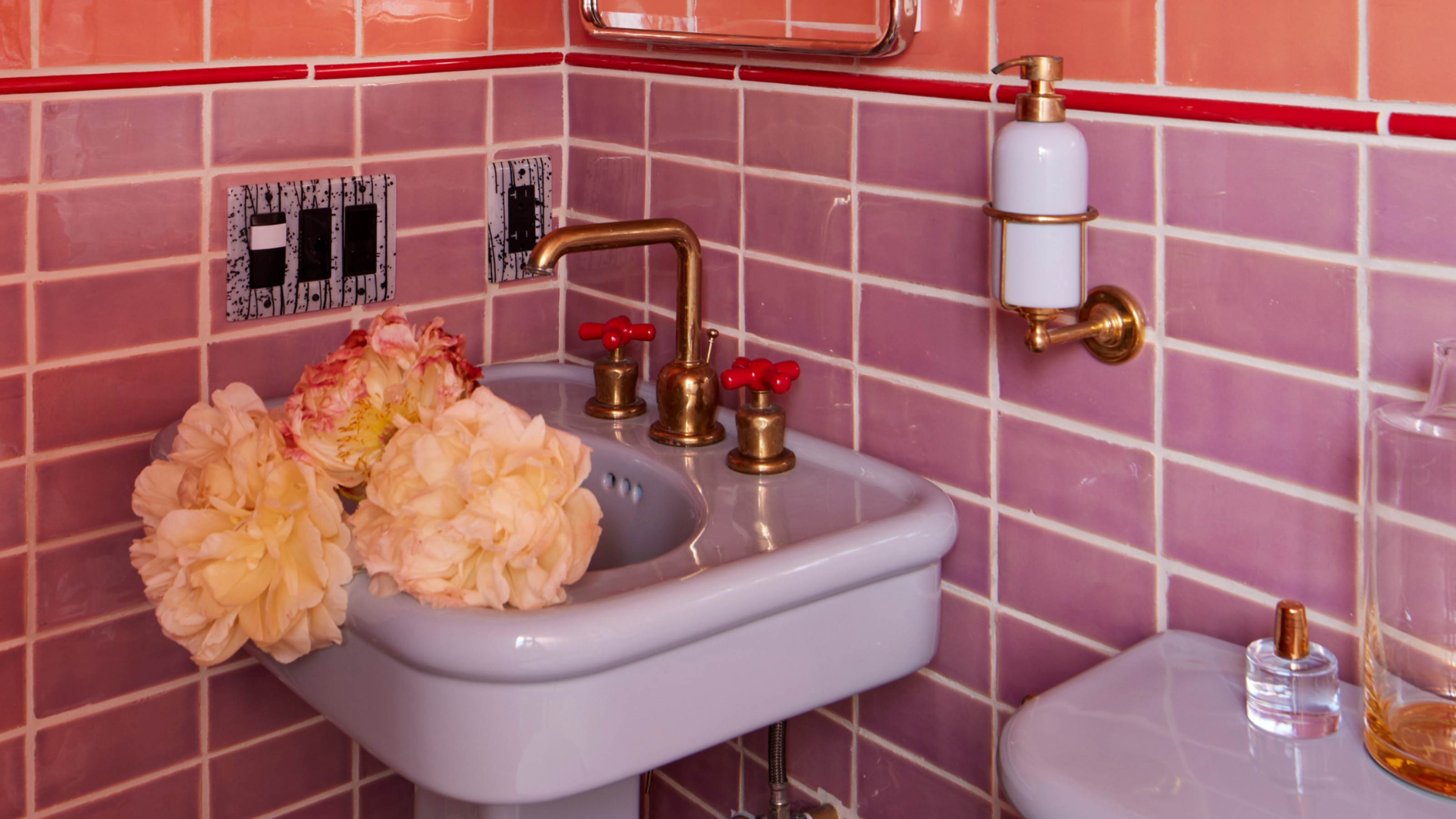 A small bathroom with dual tile drenching in a coral pink and soft purple. There is a curved mirror on the wall, cut flowers in the sink and brass hardware on the sink and the wall-attached soap dispenser