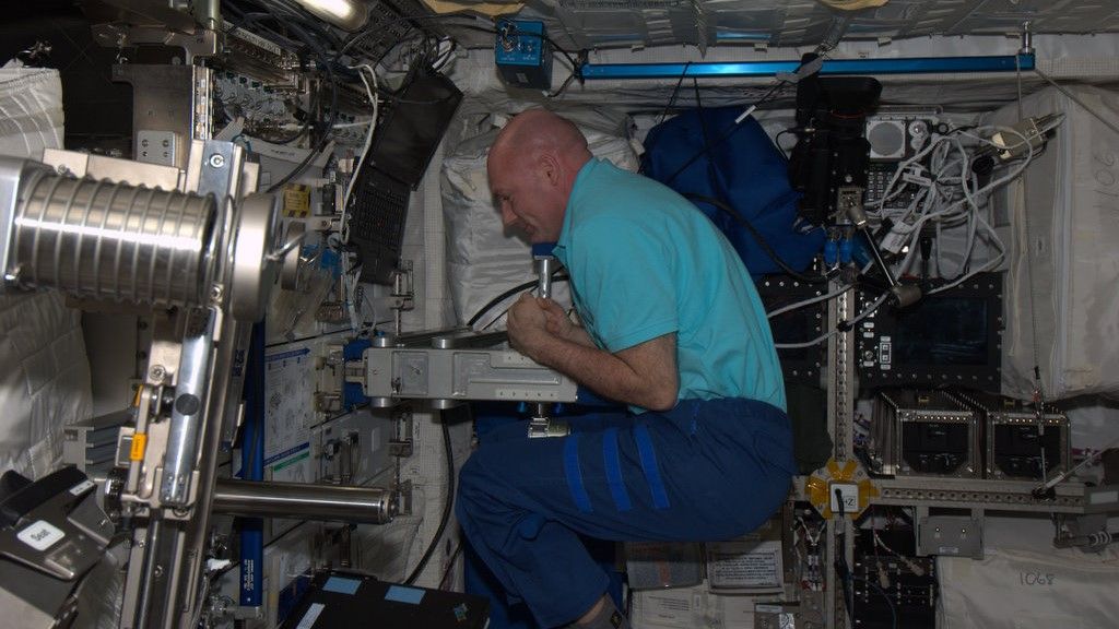 a bald man in a light blue shirt and dark blue pants scrunches his body into a ball around a white piece of metal machinery, floating in a busy spacecraft.