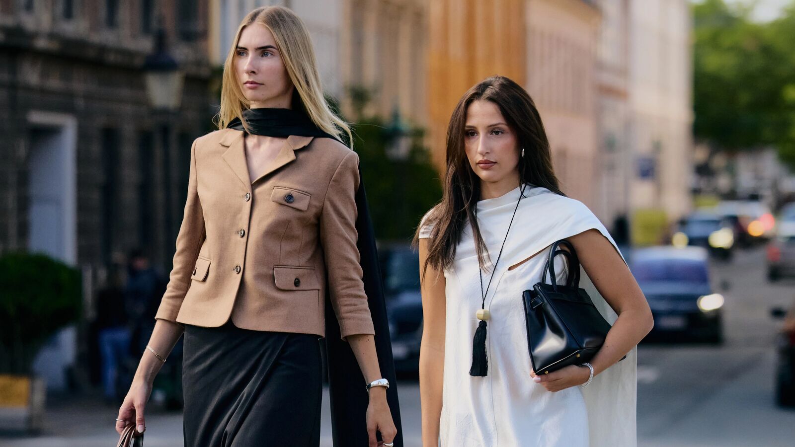 Stylish women pose in the streets of Copenhenhagen. The woman on the right wears a white dress with a cape and scarf elements with black accessories. The other woman wears a tan blazer, black scarf, black pencil skirt over trousers. 