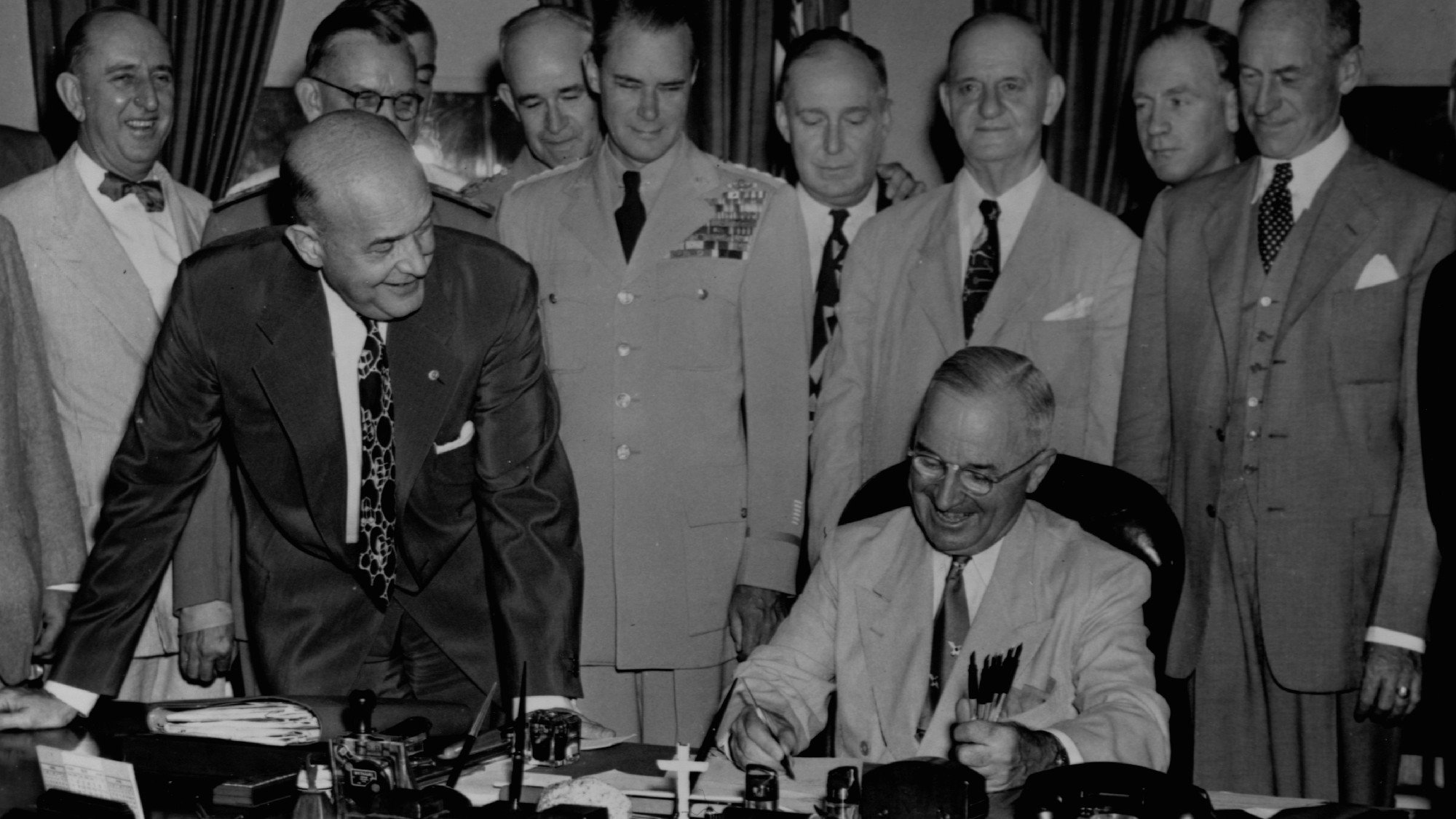 President Truman seated at a desk signing a document surrounded by political and military officials