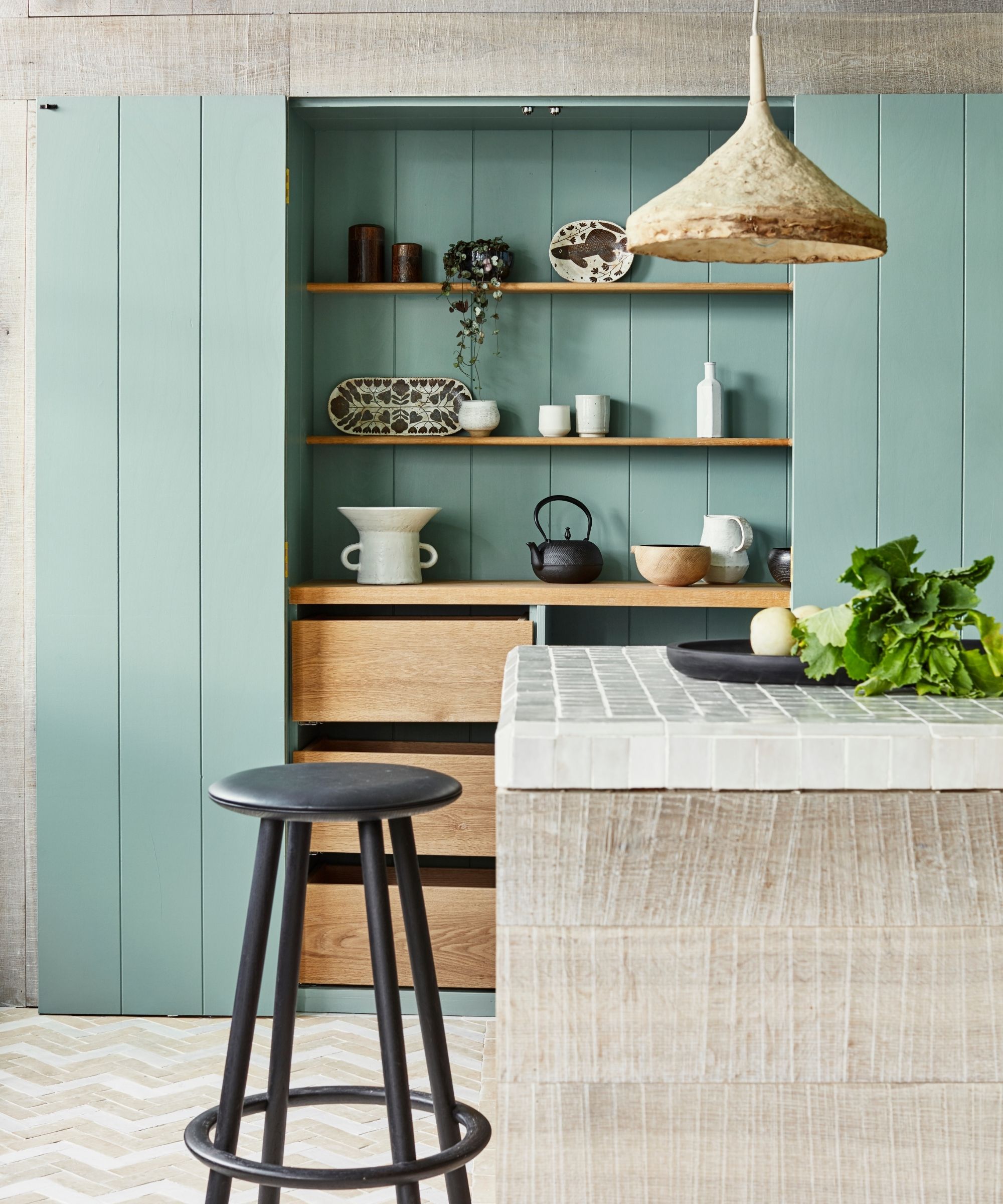 Wabi sabi kitchen with blue panelled shelving and wooden drawers, white tiled light wood kitchen island, tiled floor, and circular black stool.