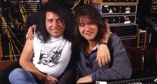 Steve Lukather and Eddie Van Halen pictured in the studio together. Lukather wears a white sleeveless T-shirt. Van Halen wears a grey longsleeve. Studio equipment can be seen in the background.