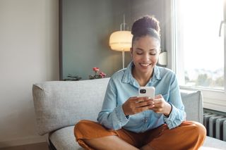 A woman sits cross-legged on her sofa looking at her smartphone and smiling