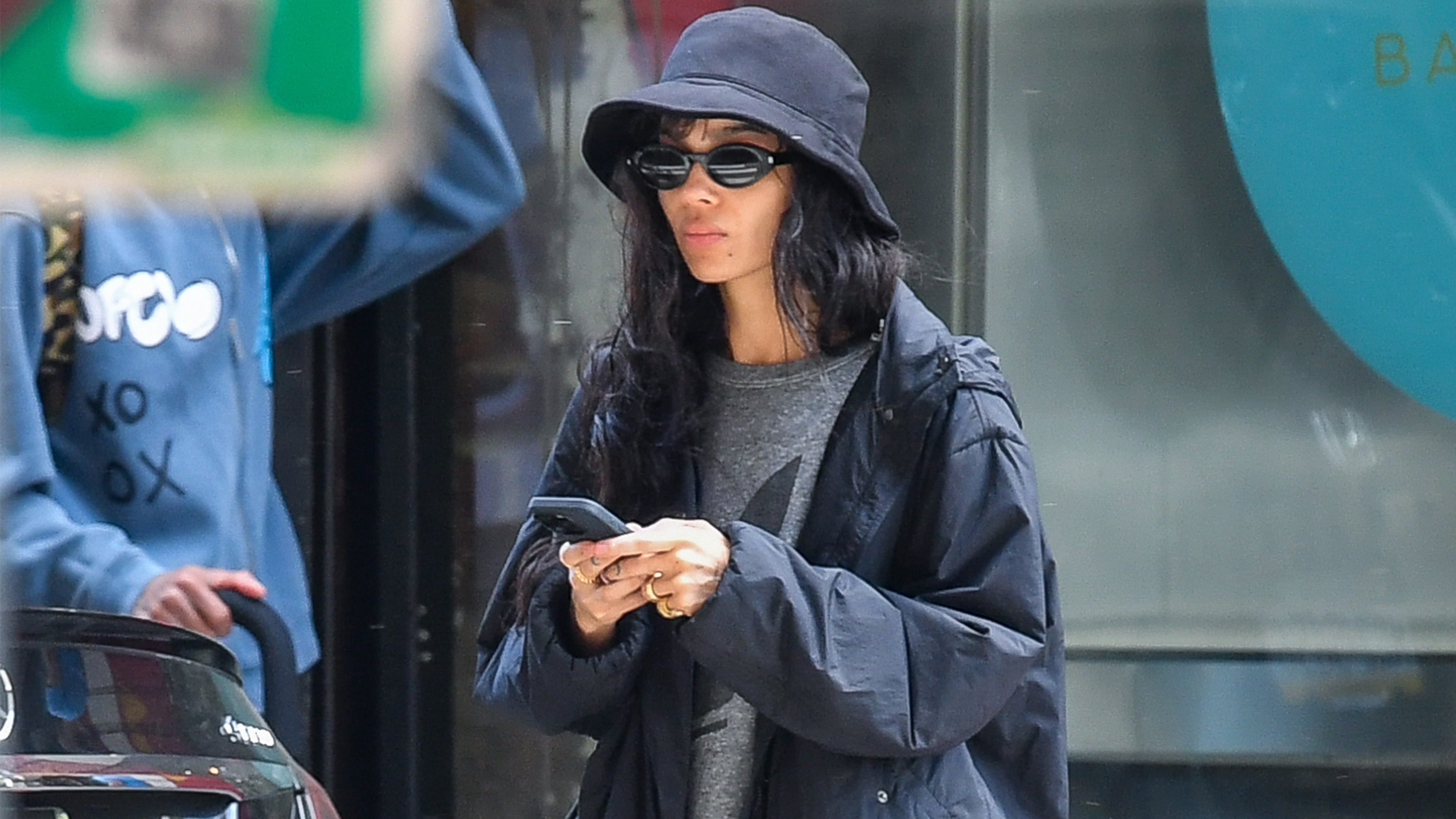 Zoe Kravitz wears a black hat, gray t shirt, black jacket, black pedal pusher leggings, and black flats.